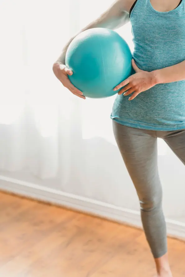 A woman in a teal tank top and gray leggings holds a turquoise exercise ball at waist height by a sunlit window—ideal for kinesiology pelvic floor active rehab in Vancouver & Kitsilano, offering exercise‑focused active rehab for core strength and chronic lower back pain relief.